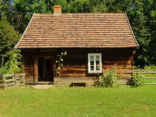 Drewniana chata z glinianym dachem, typowa dla skansenów w Polsce, otoczona zielenią i drewnianym płotem.