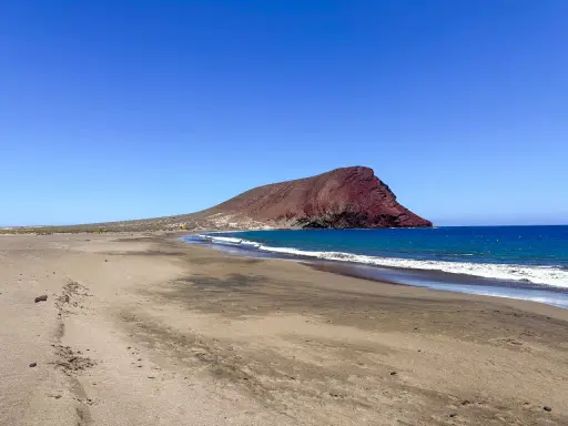 Plaże z białym piaskiem na Teneryfie, z charakterystyczną czerwoną górą w tle i turkusowym oceanem.