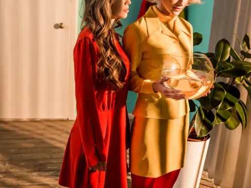 Dwie kobiety w stylizacjach w stylu retro, jedna w czerwonej sukience, druga w żółtym kostiumie, trzymająca kulę z rybkami.