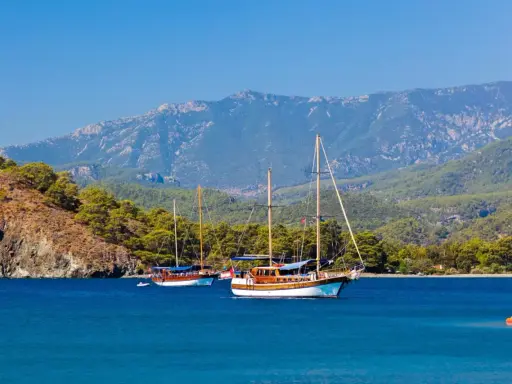 Dwa drewniane jachty kołyszą się na turkusowych wodach, zalesione wzgórza i góry w tle. Idealny dzień na zwiedzanie wybrzeża w okolicach **Antalya miasto**.