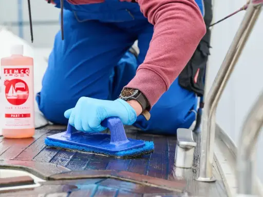 Person reinigt mit blauer Bürste und SEMCO Teak Cleaner das Holzdeck eines Bootes.
