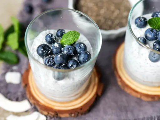 Zwei Gläser mit cremigem Chia Pudding, garniert mit Blaubeeren, Kokosraspeln und Minze. Ein gesunder Genuss!