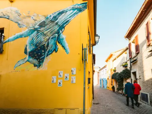Un murale di balene blu su un muro giallo a San Giuliano Rimini. Una coppia passeggia lungo la strada acciottolata.