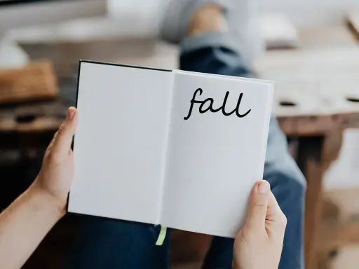 Otwarta książka z napisem "fall" na prawej stronie. Osoba w dżinsach trzyma ją, przygotowując się do jesiennych zapisków.