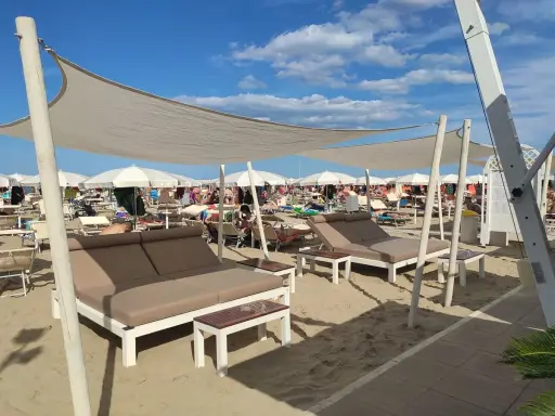Un lido a Riccione con lettini doppi e ombrelloni bianchi sotto un cielo azzurro e nuvoloso.