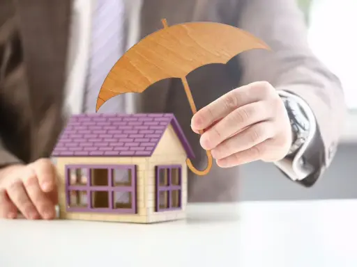 Main d'un homme tenant un parapluie en bois au-dessus d'une maison miniature. Une image symbolisant l'assurance habitation obligatoire.