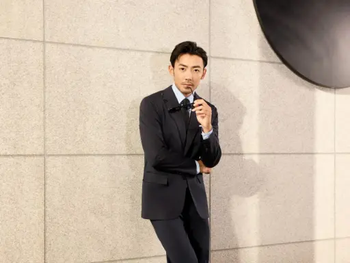 A man in a black suit and black shirt, holding sunglasses, leans against a textured wall.