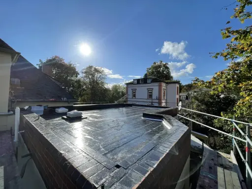 Ein frisch abgedichtetes Flachdach unter strahlender Sonne. Die Dachhaut glänzt, zwei Dachfenster sind sichtbar.