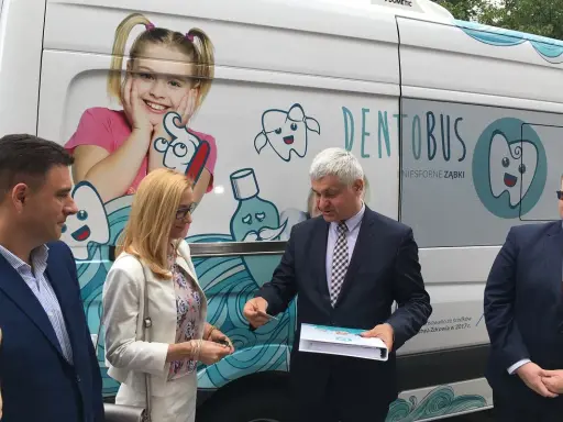 Ludzie stojący przed białym Dentobusem z grafikami uśmiechniętych zębów i dziewczynki.
