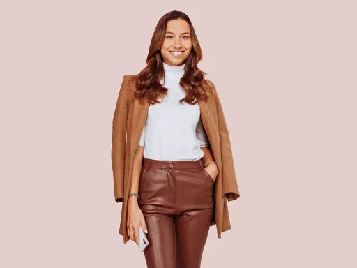 Donna sorridente con un elegante outfit con pantaloni di pelle larghi marroni, dolcevita bianco e cappotto cammello.