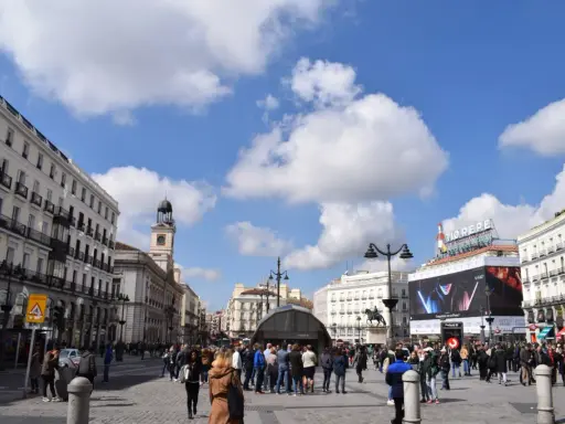 Menschenmassen beleben den Platz Puerta del Sol in Madrid unter einem blauen Himmel mit Wolken. Der Uhrturm und das Tio Pepe Schild sind sichtbar.