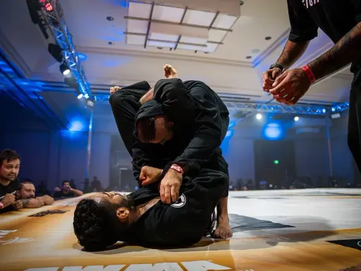 Dwóch mężczyzn w czarnych kimonach walczy w parterze. Intensywna walka jiu jitsu, z widownią w tle.