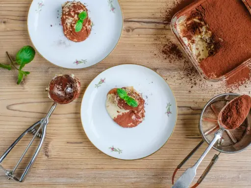 Tiramisu anrichten: Ein Löffel voll cremigen Tiramisus, bestäubt mit Kakao, wartet auf seinen Einsatz. Daneben Teller mit dem fertigen Dessert, garniert mit Minze.