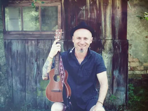 Uśmiechnięty mężczyzna w kapeluszu trzyma ukulele, gotowy do grania nim zajdzie słońce.
