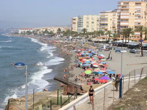 Sonniger Torrox Strand mit vielen bunten Sonnenschirmen, Menschen im Meer und an Land, sowie historischen Kanonen.