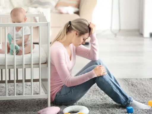 Zmartwiona mama siedzi obok łóżeczka, zastanawiając się, depresja poporodowa ile trwa. Dziecko bawi się w łóżeczku.