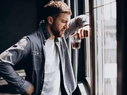 Mężczyzna zamyślony przy oknie, trzymający szklankę z bursztynowym płynem. Czy to jeden z ukryty alkoholizm objawy?