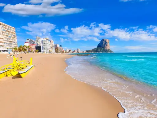 Sonne, Strand und Meer in Calpe, Spanien. Ein gelbes Wasserfahrrad wartet am Sandstrand, im Hintergrund die Stadt und der markante Felsen Peñón de Ifach. Ein echter Geheimtipp!
