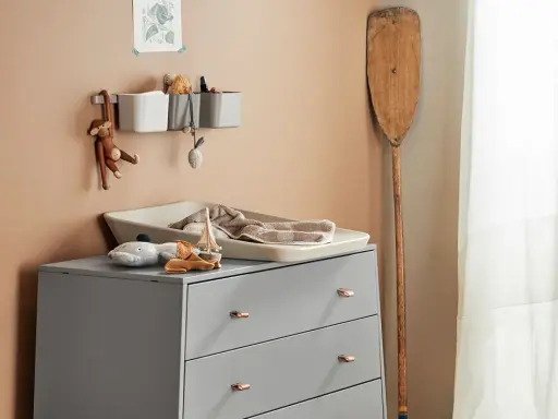 Eine Wickelkommode organisieren: Spielzeug, Handtücher und Pflegeutensilien sind ordentlich verstaut. Ein Holzpaddel lehnt an der Wand.