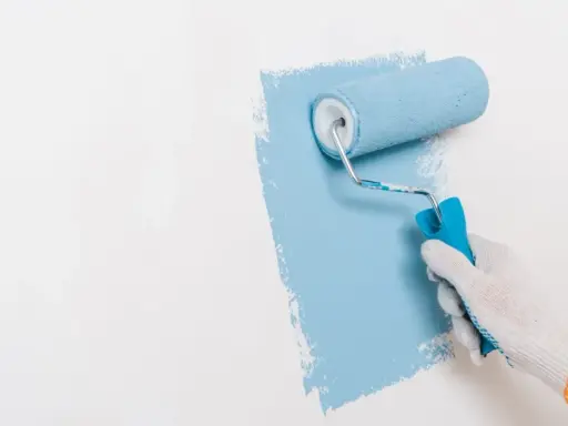 Ręka w białej rękawiczce maluje ścianę na niebiesko wałkiem. Widać nierówności, co może sugerować wady farby ceramicznej.