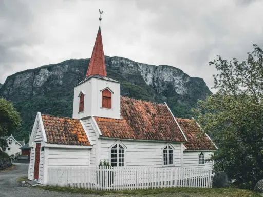 Biały kościółek z czerwoną wieżą i dachem, otoczony białym płotkiem, na tle surowych gór. Symbol norweskiej religii i spokoju.