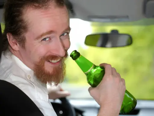 Uśmiechnięty mężczyzna z rudą brodą trzyma zieloną butelkę piwa w samochodzie. Mówi, że piwo to nie alkohol, ale jazda z nim jest niebezpieczna.