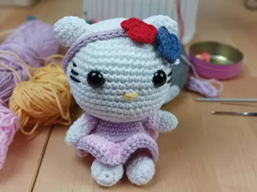 Muñeca de Hello Kitty tejida a crochet, con vestido lila y lazo rojo y azul. Hilos y agujas de crochet alrededor, perfectos para crear tus propios hello kitty crochet patrones español.
