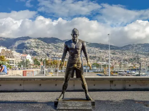 Bronzestatue von Cristiano Ronaldo am Hafen von Funchal, Madeira. Hinter ihm erstreckt sich die Stadt bis zu den Bergen. Ein Denkmal für den weltberühmten Fußballer, das man fast wie am **cristiano ronaldo flughafen** erwarten könnte.