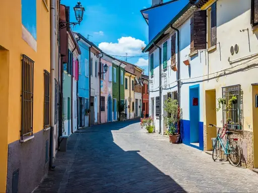 Un vicolo colorato a San Giuliano, con case dipinte di giallo, blu, verde e rosa. Una bicicletta azzurra è parcheggiata davanti a una casa.