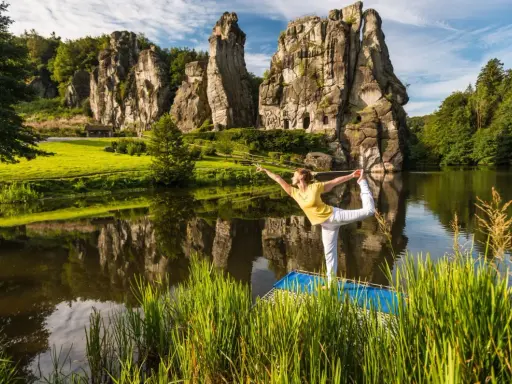 Frau praktiziert Yoga am See vor den Externsteinen NRW. Die Felsen spiegeln sich im Wasser.