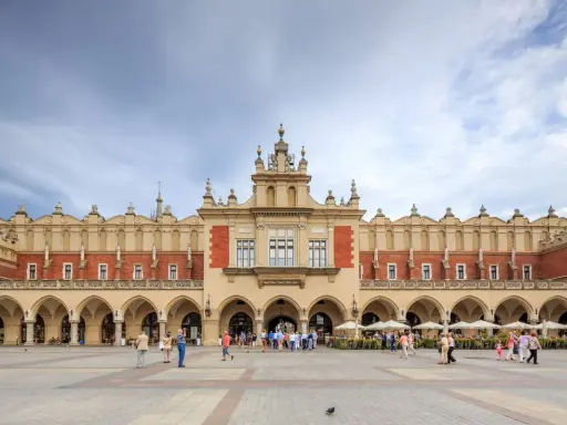 Sukiennice w Krakowie, zabytkowy budynek z arkadami, tętni życiem. Ludzie spacerują po Rynku Głównym, podziwiając architekturę.