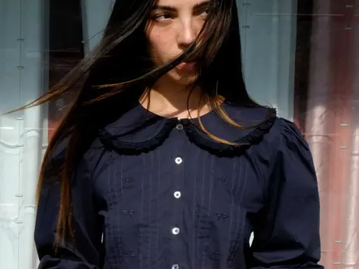 A young woman with windswept hair wears a dark blue blouse with a charming peter pan neckline, delicate embroidery, and puffed sleeves.