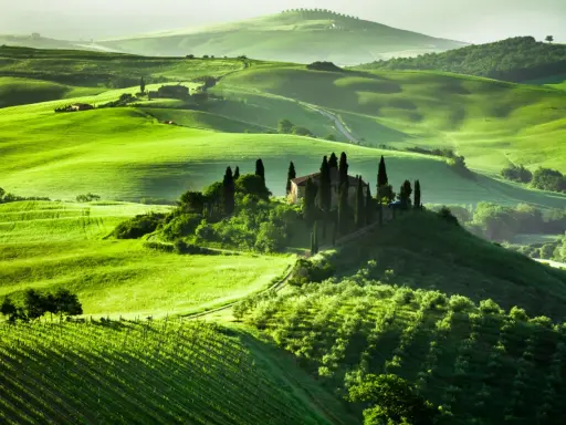 Colline verdi ondulate in Toscana, con una villa su una collina circondata da cipressi. Un sogno per un viaggio in Toscana.