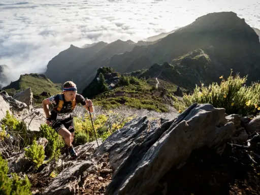 Läufer kämpft sich auf Madeira bergauf, vorbei an Felsen und gelben Ginsterbüschen, mit Wolkenmeeren und schroffen Gipfeln im Hintergrund.