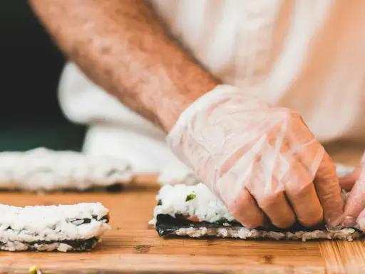 Hände mit Handschuhen rollen Sushi auf einem Holzbrett. So einfach ist Sushi selbst machen!