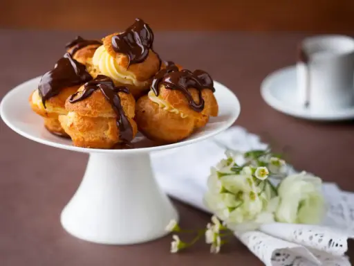 Un plateau blanc présente des profiteroles gastronomiques garnies de crème et nappées de chocolat noir. Des fleurs délicates ajoutent une touche de fraîcheur.