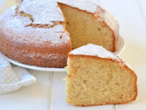 Una fetta di torta di banane soffice, cosparsa di zucchero a velo, accanto alla torta intera su un piatto bianco.