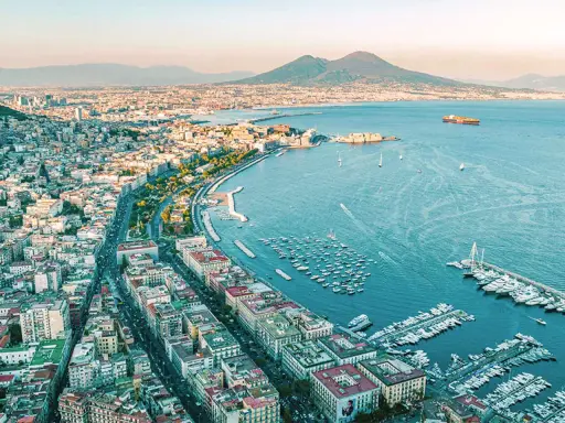 Vy över Neapel med Vesuvius i bakgrunden. Hamnen är full av båtar, och staden sträcker sig längs kusten. En perfekt vy innan man flyger från neapel flygplats.