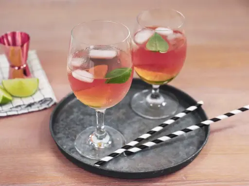 Zwei Gläser Rosato Spritz mit Eis und Minze, dazu Limettenspalten und ein Messbecher. Perfekt für einen sommerlichen Genuss.