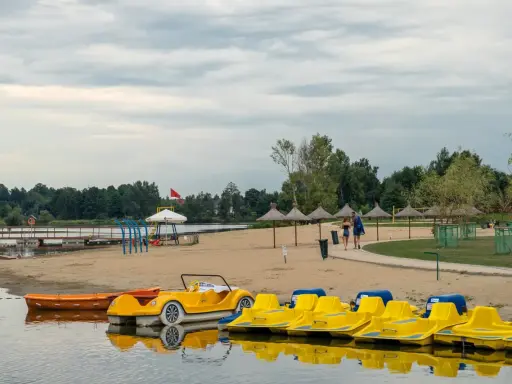 Żółte rowerki wodne i łódki czekają na plaży nad jeziorem blisko Warszawy. Para spaceruje po piasku pod słomianymi parasolami.