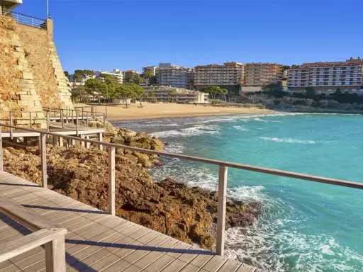 Paseo marítimo de madera junto a rocas y mar turquesa, con playa y edificios al fondo. Un tramo del camí de ronda Tarragona.