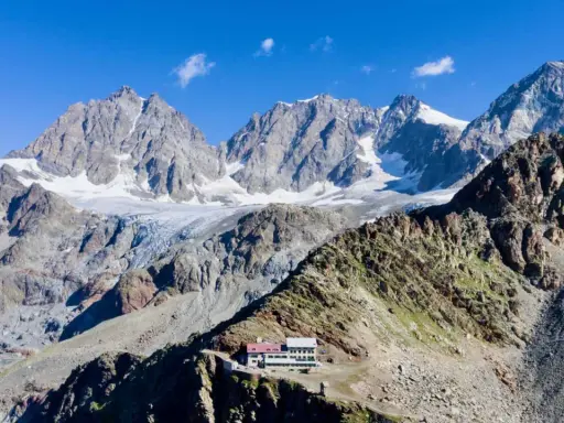Il rifugio Marinelli, un edificio bianco e rosso, si erge su una cresta rocciosa con imponenti montagne innevate sullo sfondo.
