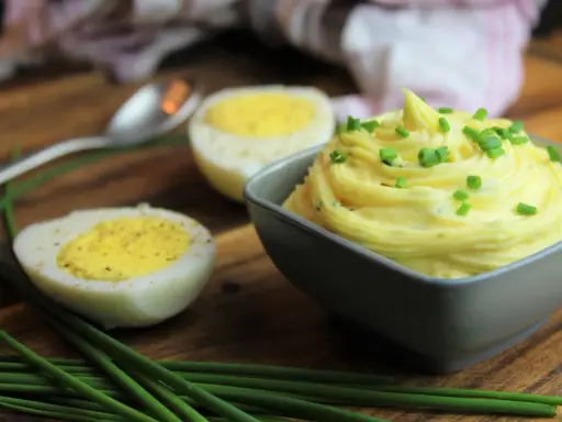 Cremige Sauce Tartar, garniert mit Schnittlauch, neben halbierten Eiern auf einem Holzbrett.
