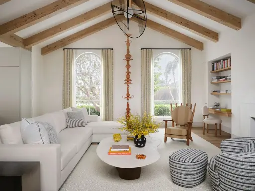 Salón luminoso con vigas de madera en el techo, creando la sensación de casas con techos bajos. Sofá blanco, mesa ovalada y butaca a rayas.
