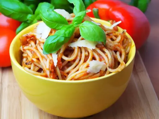 Pyszne spaghetti bez mięsa w żółtej misce, udekorowane bazylią i parmezanem. Obok pomidory i papryka.
