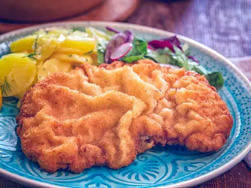 Ein knuspriges, originales Wiener Schnitzel auf einem blauen Teller, serviert mit Kartoffelsalat und frischem Grün.