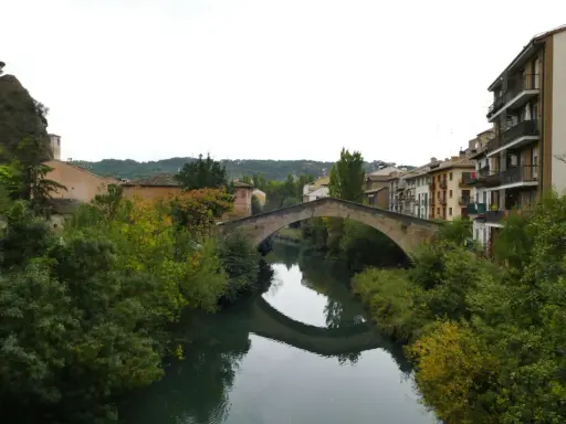 El puente de la cárcel de Estella cruza un río sereno, reflejando su arco de piedra entre frondosa vegetación y edificios históricos.