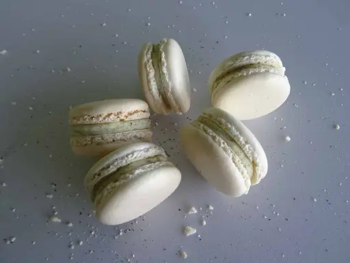 Cinq macarons vanille, délicatement posés sur une surface grise parsemée de miettes.