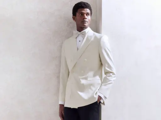 A Black tie wedding guest man in a cream double-breasted tuxedo with a matching bow tie, looking dapper.
