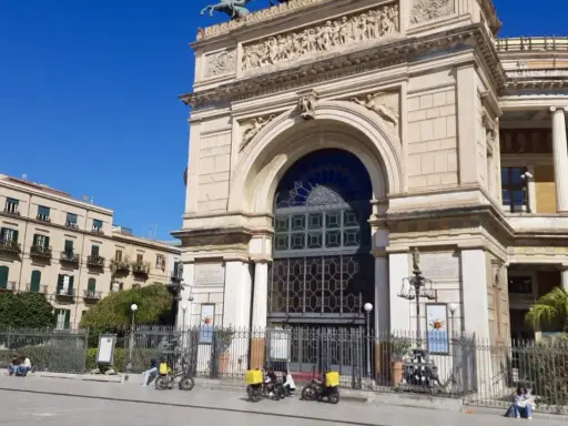 Il **Teatro Politeama Garibaldi** di Palermo, con la sua imponente facciata neoclassica, si staglia sotto un cielo azzurro. Tre scooter elettrici gialli sono parcheggiati davanti.